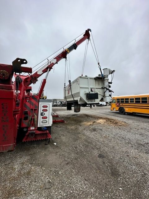 A large, white electrical transformer being lifted by a red tow truck in an outdoor lot, a yellow school bus is in the background.