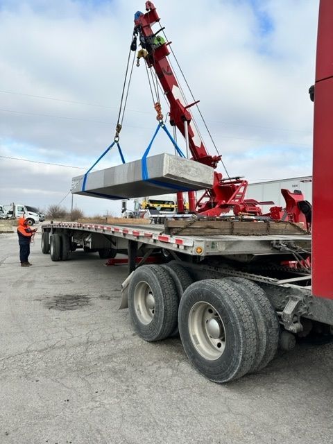 A red crane lifts a large, rectangular concrete slab onto a flatbed trailer. A worker in an orange vest stands nearby.