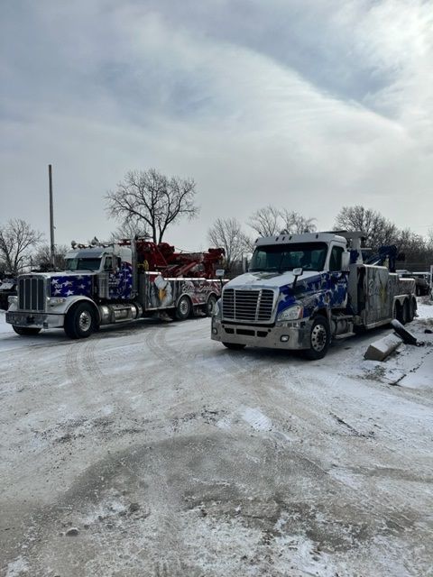 Two tow trucks parked on a snow-covered lot on a cloudy day. The trucks have blue and silver graphics.
