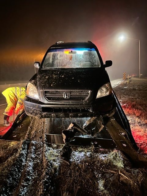 Black SUV crashed, front end damaged, on a muddy road at night with a person in a yellow jacket inspecting the damage.