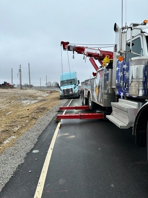 A tow truck lifting a light blue semi-truck out of a ditch on a gray, overcast day. The tow truck is on a paved road.