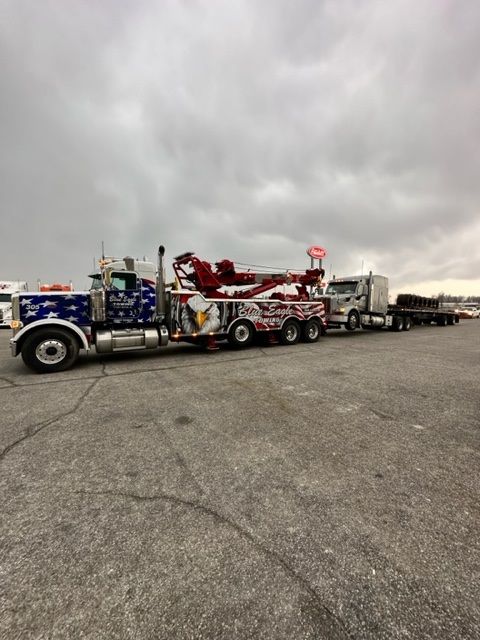 A large tow truck with patriotic designs is towing a semi-truck and trailer on a gray, cloudy day.
