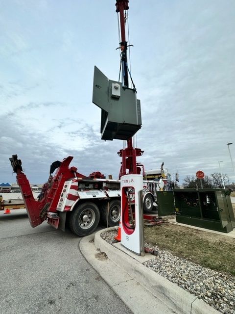 A tow truck is lifting a large electrical component above a Tesla charging station on a cloudy day.