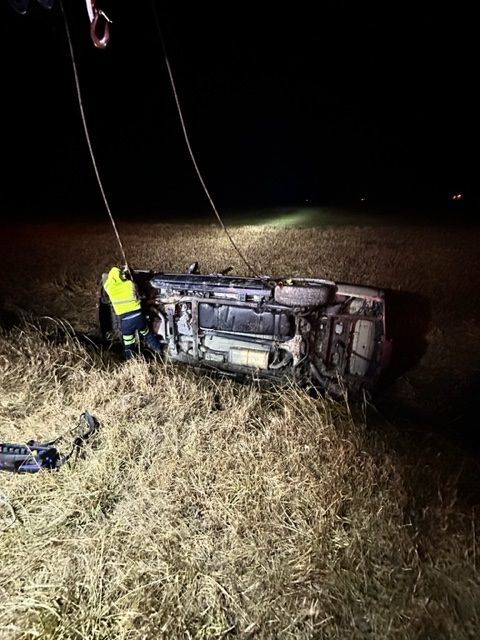 A vehicle overturned in a field at night, with a tow truck preparing to upright it. A person in a yellow vest is present.