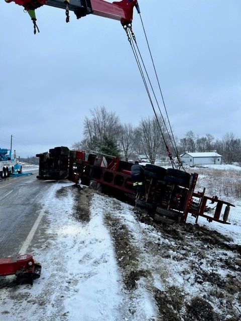 A red overturned semi-truck being lifted by a tow truck on a snow-covered roadside. The sky is overcast.