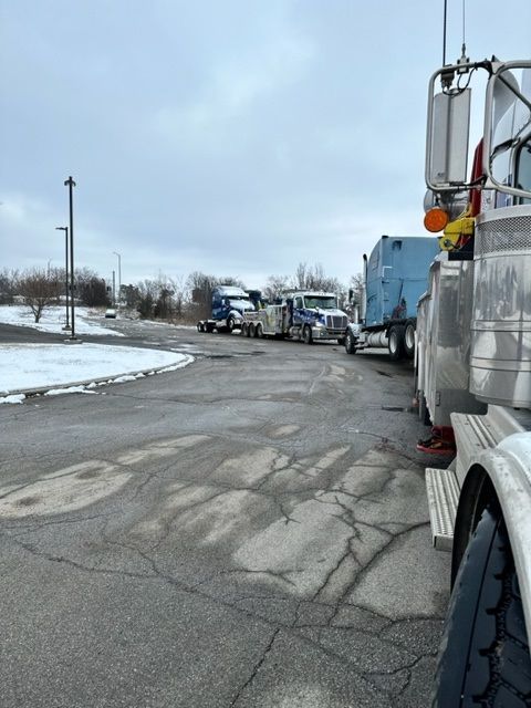 Semi-trucks parked in a lot on a cloudy day, some in front of others. Snow on the ground, asphalt in foreground.