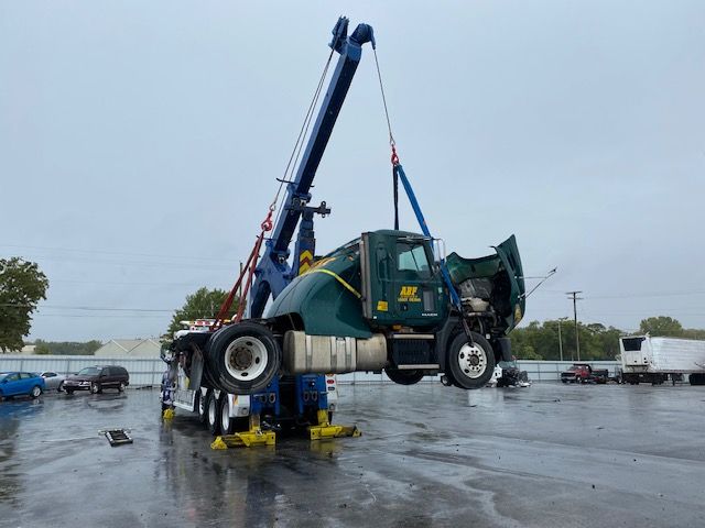 A tow truck lifting a green semi-truck cab in a wet, overcast lot. The cab's hood is open, and the tires are off the ground.