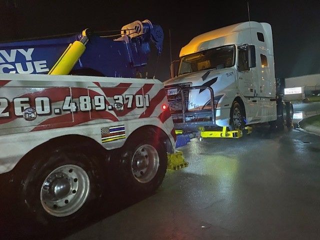 A tow truck is towing a damaged white semi-truck at night. The tow truck has blue and white markings.
