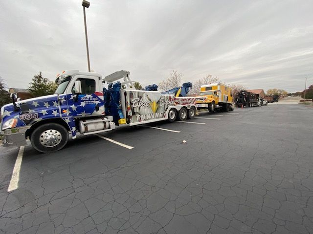 Tow truck with American flag design towing vehicles in a parking lot on an overcast day. Other tow trucks line up behind.