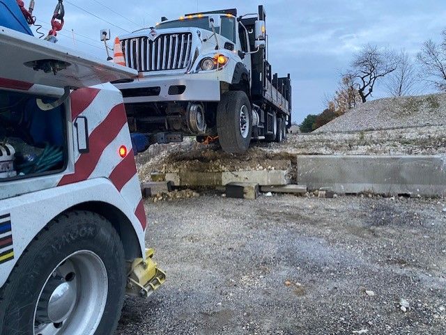 Tow truck attempting to pull a large white semi-truck off of a concrete barrier in a gravel area.