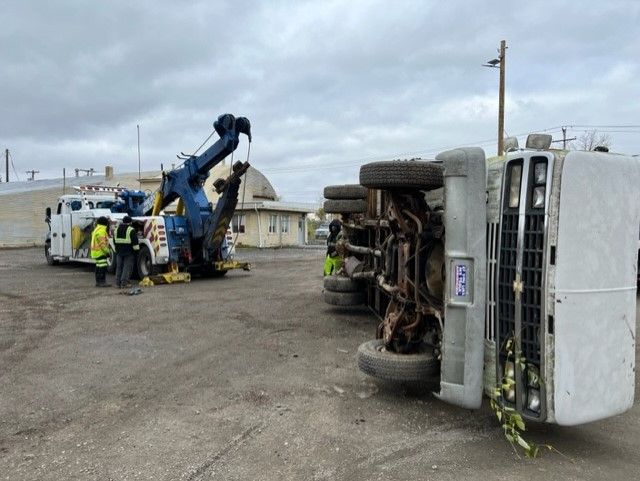 A tow truck attempts to upright a semi-truck that has overturned. Two workers in safety vests stand nearby on a gray asphalt surface.