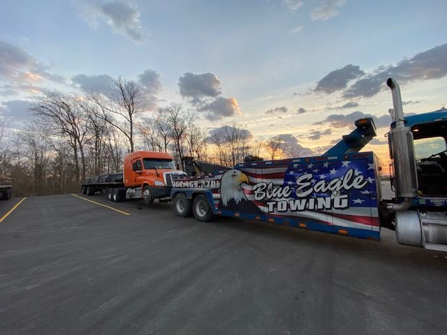 A blue tow truck with patriotic graphics tows an orange semi-truck on an asphalt lot at sunset.