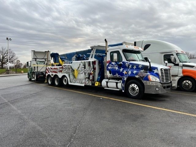A patriotic tow truck hauling debris at a parking lot, next to another semi-truck, under a cloudy sky.