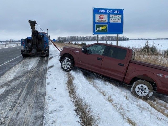 A maroon pickup truck stuck off a snow-covered road is being towed. A blue tow truck is attached. 