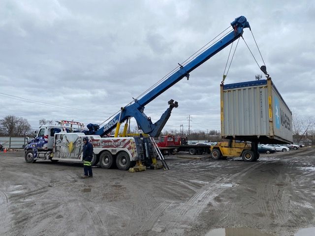 A large blue tow truck is lifting a shipping container in a muddy lot under a cloudy sky.