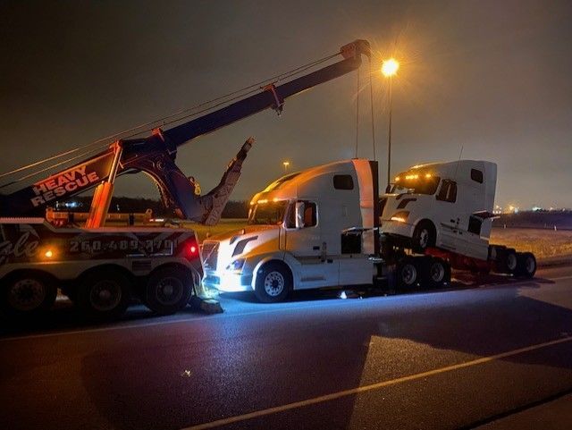 Heavy-duty tow truck rescuing two semi-truck cabs on a road at night. The tow truck has a large boom and bright headlights.