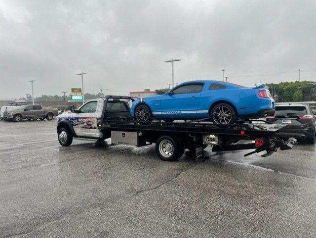 A blue Ford Mustang being towed on a flatbed tow truck on a wet, overcast day in a parking lot.