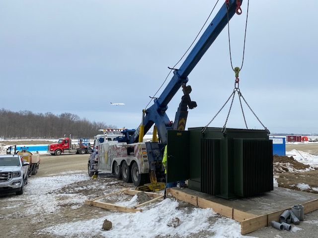 A tow truck crane lifts a large green electrical transformer at an outdoor construction site with other vehicles and a cloudy sky.