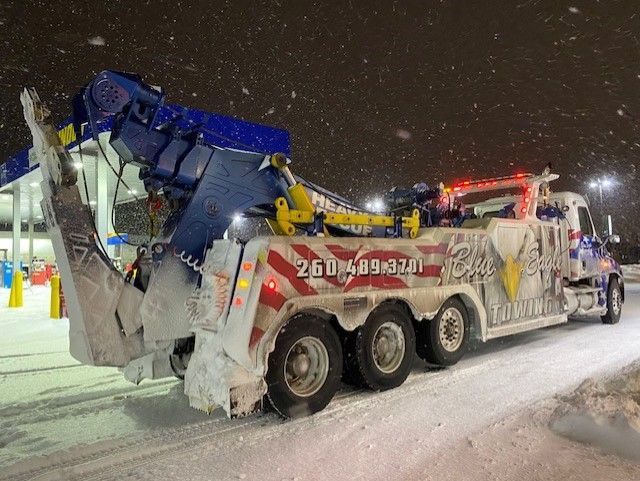A tow truck covered in snow at night, lights on. Red and blue colors on the vehicle, phone number visible.