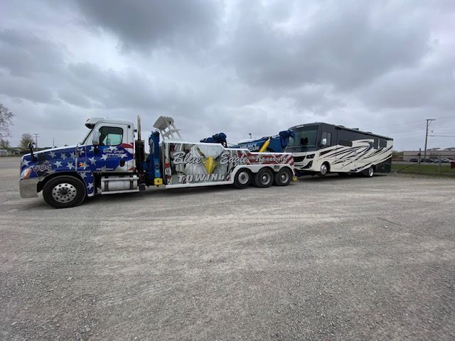 Tow truck with patriotic designs towing a recreational vehicle on a gray day. The scene appears to be a parking lot.
