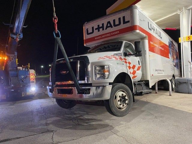 A U-Haul truck being towed by a tow truck at a gas station at night. The truck is white with red accents.