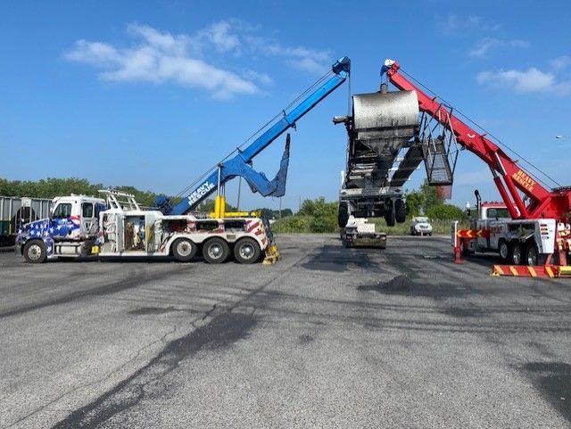 Two tow trucks lifting a burned tanker truck against a blue sky. The blue truck on the left and red on the right.