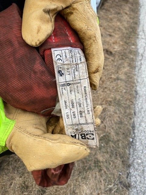 Hands wearing tan work gloves holding a red sling with a white label showing lifting capacity information.