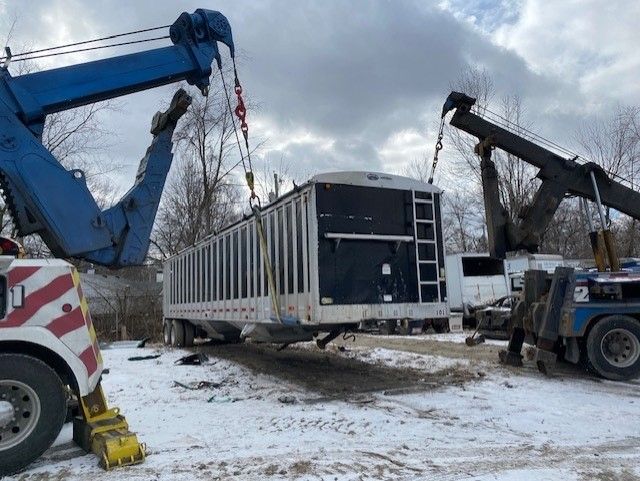 Two tow trucks lift a large white container in a snowy outdoor setting. The container has a ladder and dark panel.