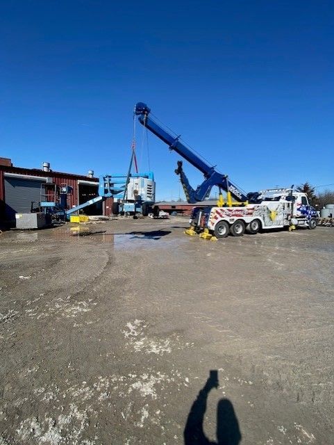 A large blue tow truck lifts a blue scissor lift with a crane on a sunny day outside a building.