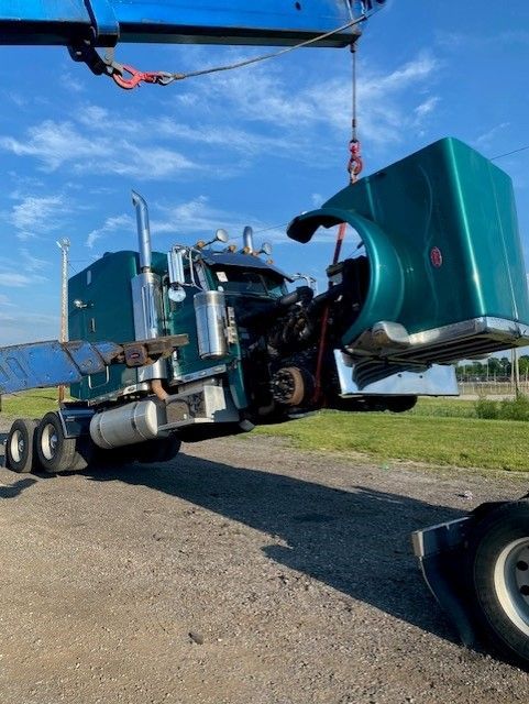A teal semi-truck with its hood raised by a crane, likely for repairs. The setting is outdoors, with a clear sky.