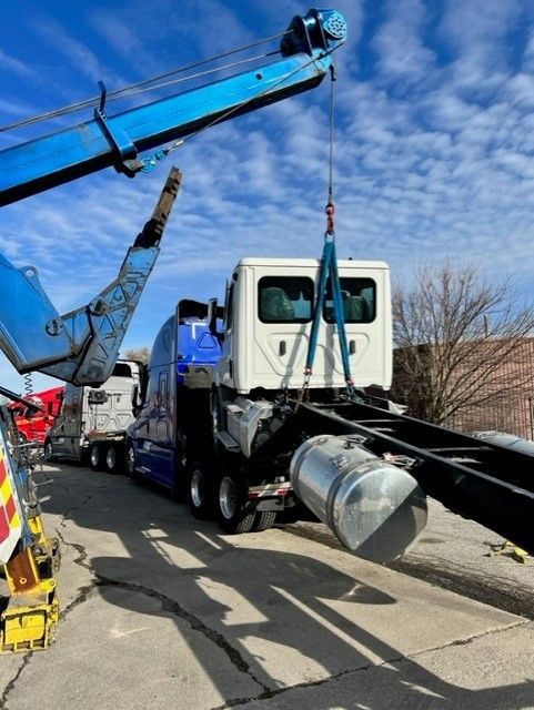 A tow truck lifts the cab of a blue and white semi-truck, likely for repair, outside on a sunny day.