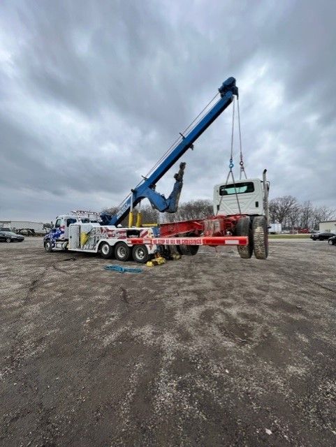 Tow truck lifting a white truck cab in a gravel lot under a cloudy sky. The tow truck is blue, red, and white.