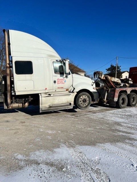 White semi-truck towing a flatbed tow truck on a snowy, asphalt surface under a clear, blue sky.