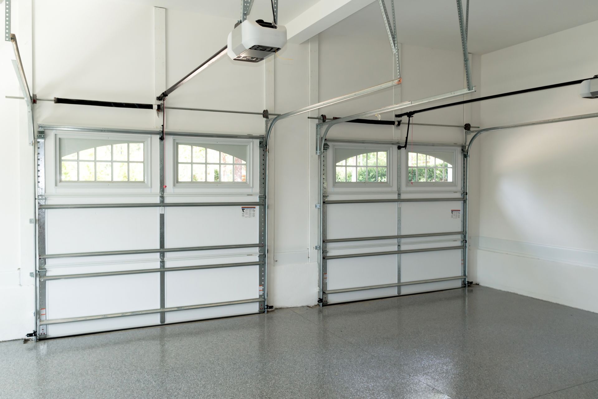 Two closed white garage doors with arched windows in a well-lit, empty garage with a speckled gray floor.