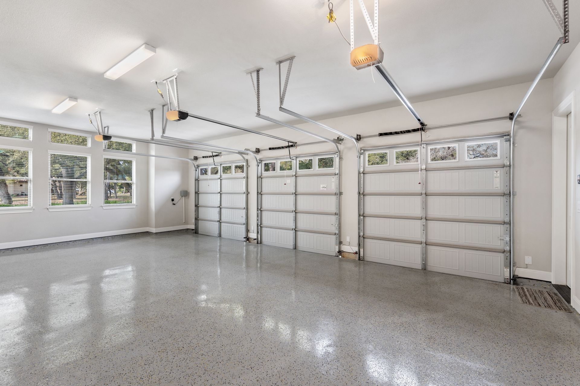 Empty three-car garage with three closed overhead doors, a glossy gray floor, and windows.