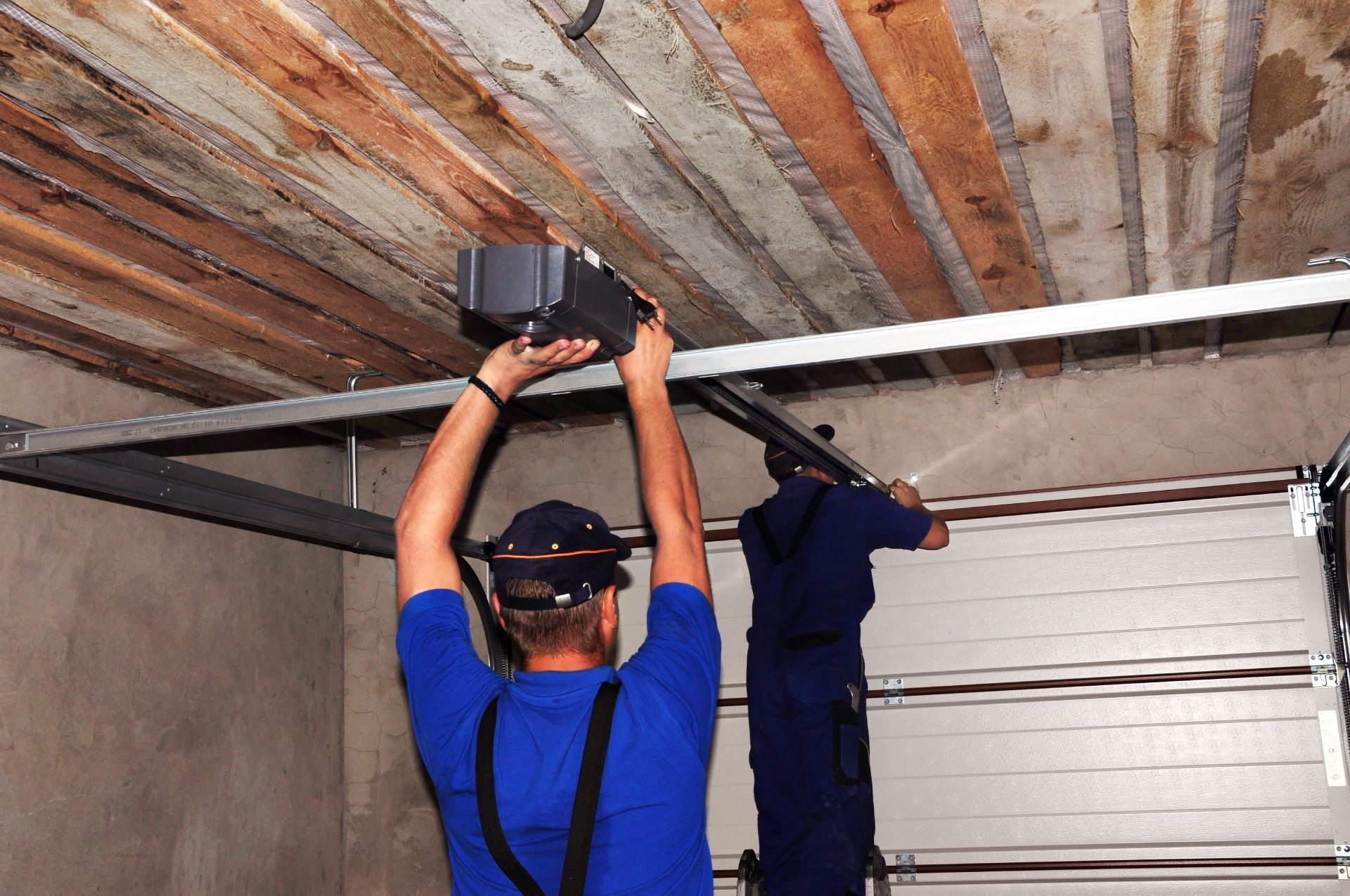 Two workers install a garage door opener on a metal track attached to a wooden ceiling.
