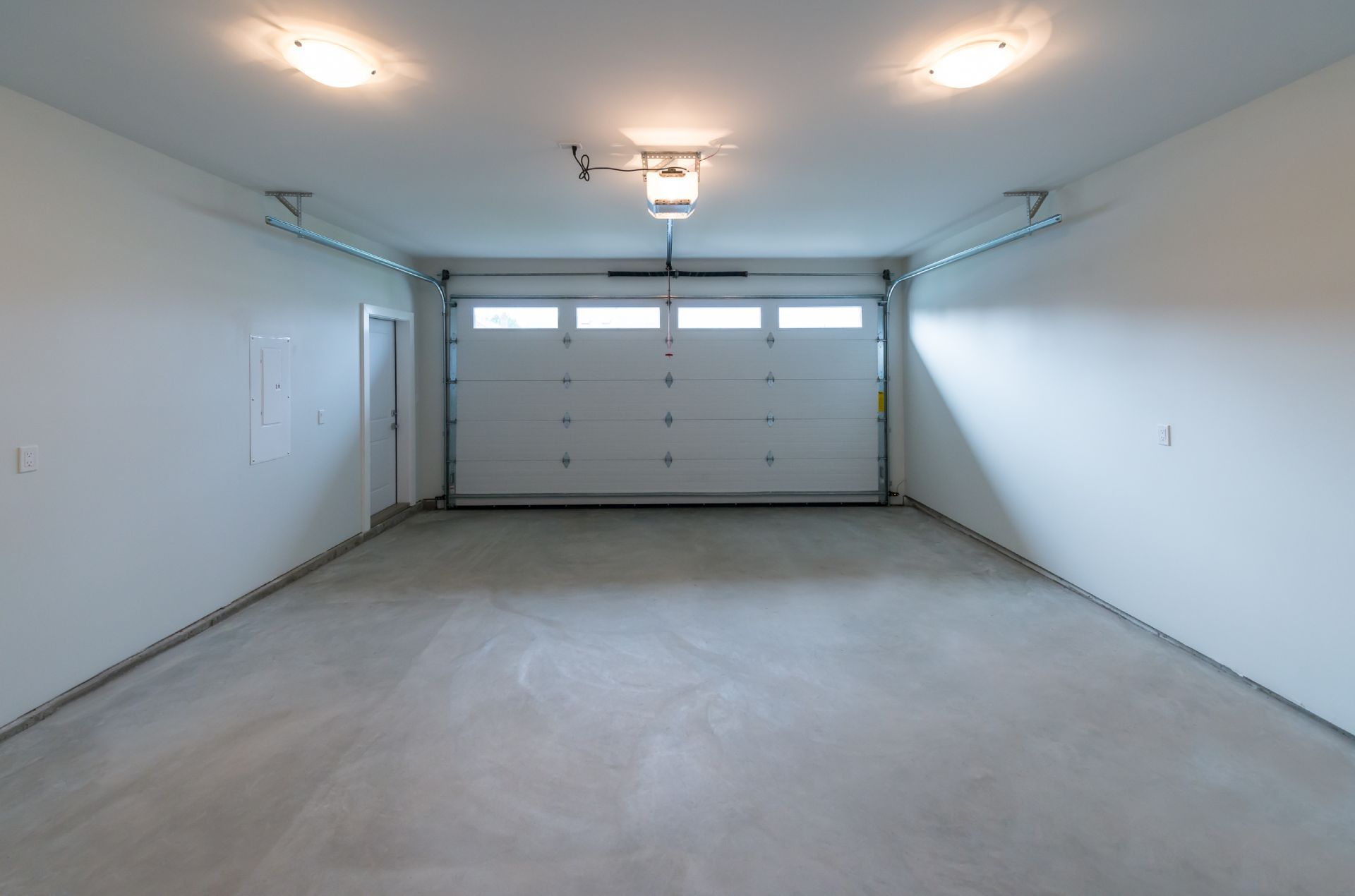 Empty, well-lit garage with closed white garage door, concrete floor, and white walls.