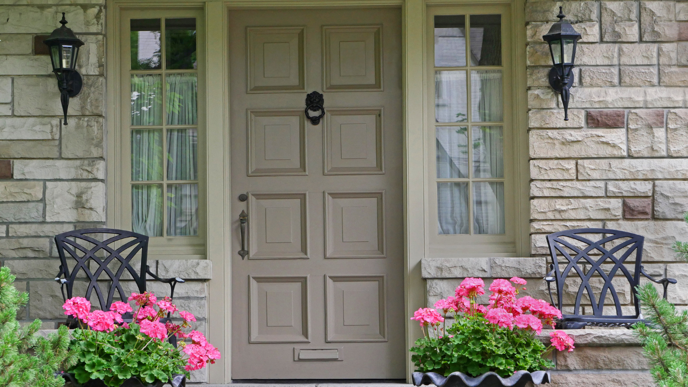 Stone exterior with tan door, two windows, and black lanterns. Pink flowers flank the doorway with two black chairs.