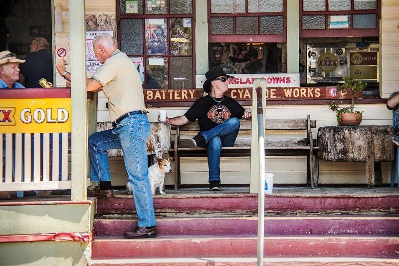 Two men are sitting on a bench with a dog in front of a building that says battery — G & D Ross Bus Charters In Magnolia, QLD
