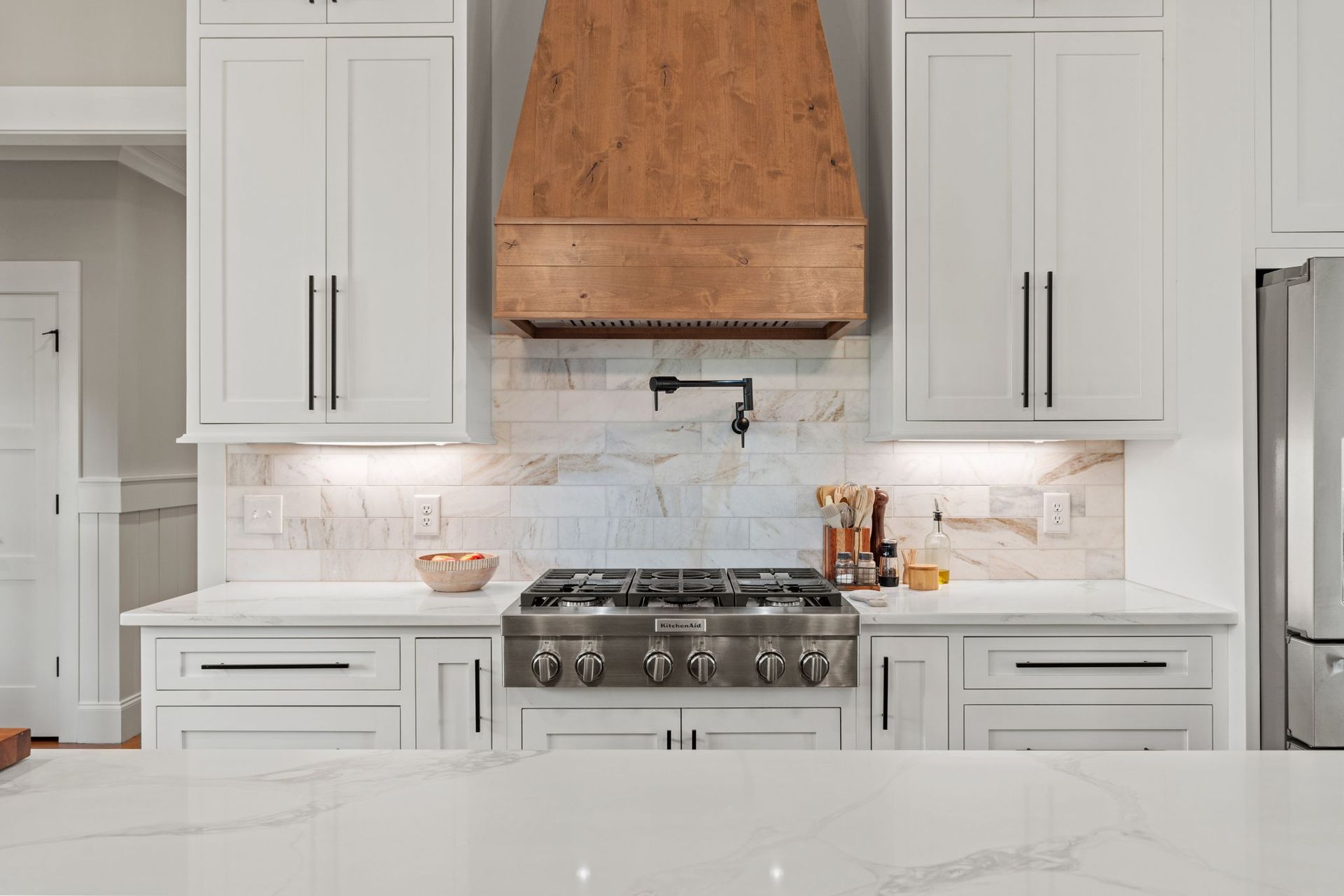 A kitchen with white cabinets , a stove , a refrigerator , and a wooden hood.