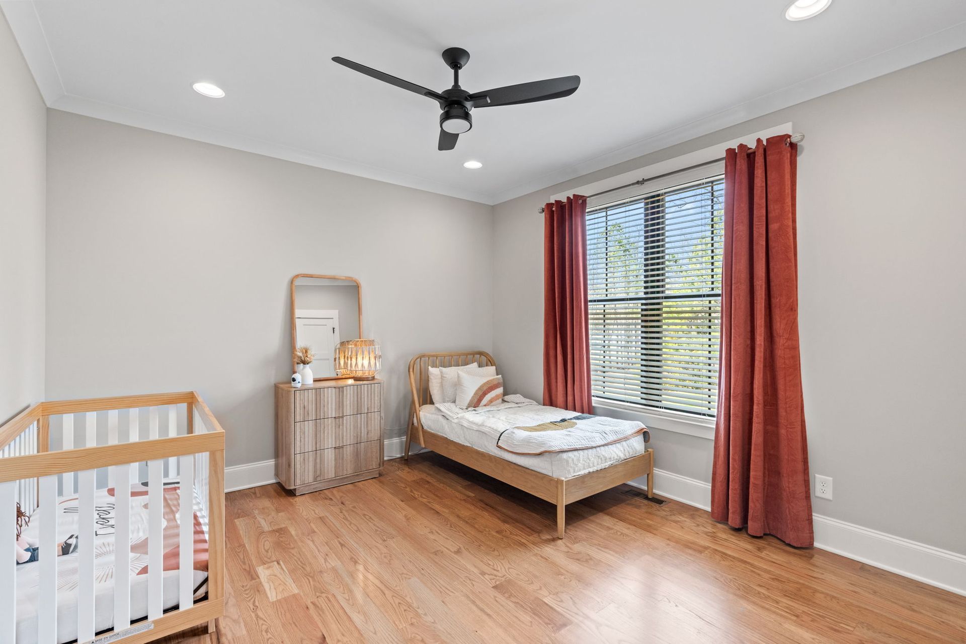 A bedroom with a bed , crib , dresser , mirror and ceiling fan.