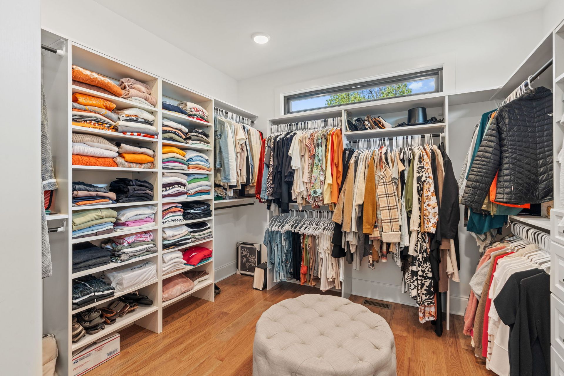 A walk in closet filled with lots of clothes and an ottoman.