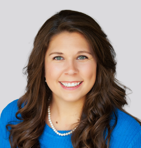A woman wearing a blue shirt and a pearl necklace is smiling.