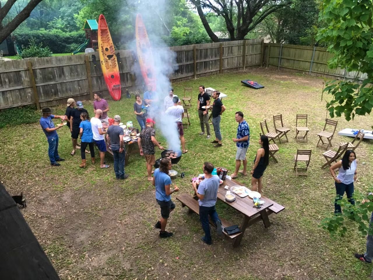 People gathered in a backyard around a smoking fire pit, with a long table and scattered chairs.