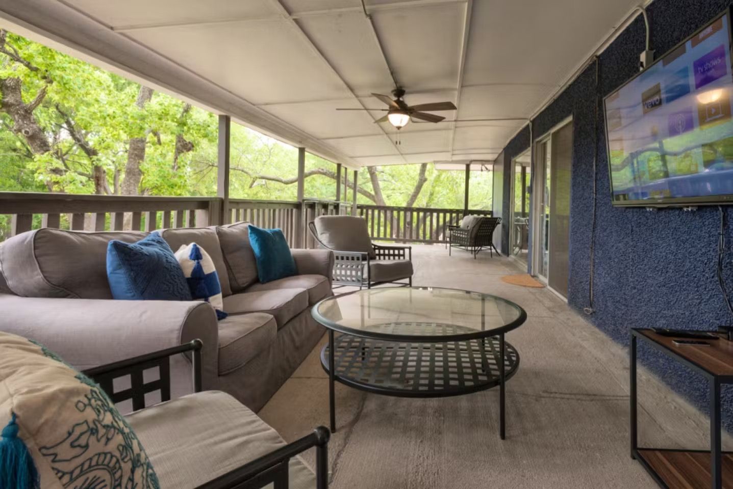 Covered patio seating area with sofa, chairs, glass coffee table, and TV overlooking greenery