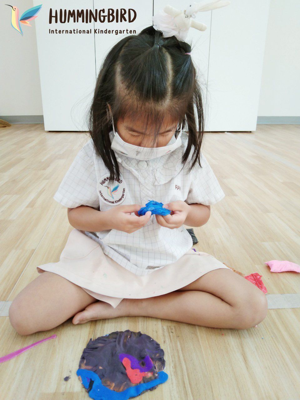 A little girl wearing a mask is sitting on the floor playing with clay.