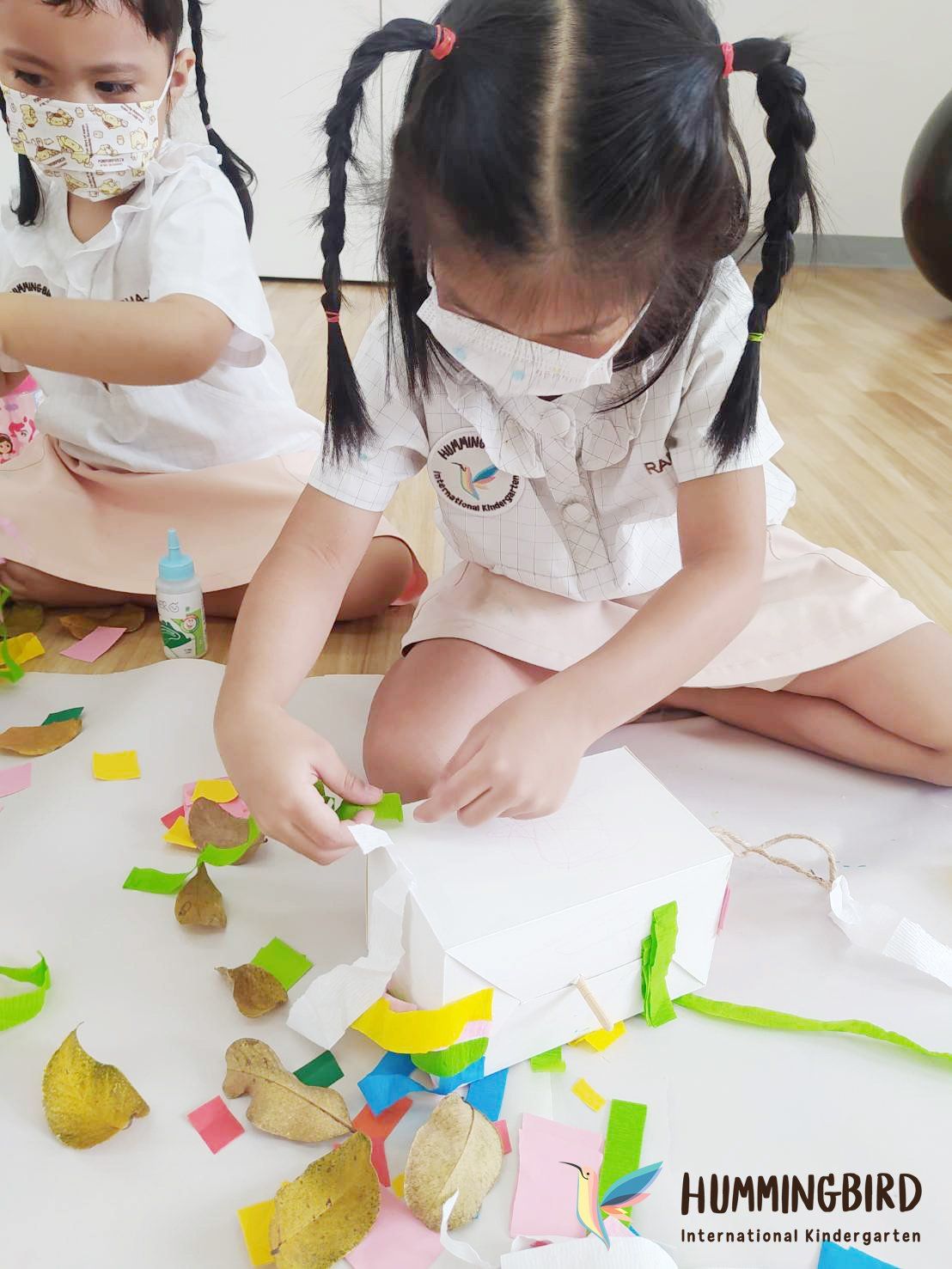 Two young girls wearing face masks are sitting on the floor making crafts.