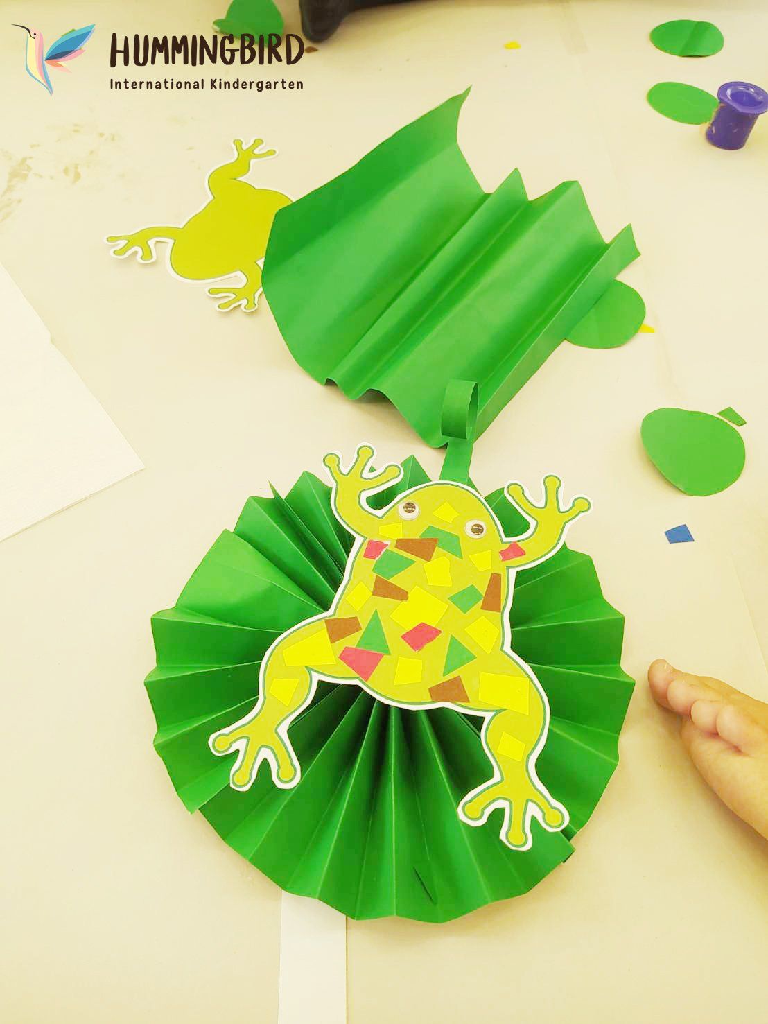 A child is making a paper frog with a hummingbird logo in the background