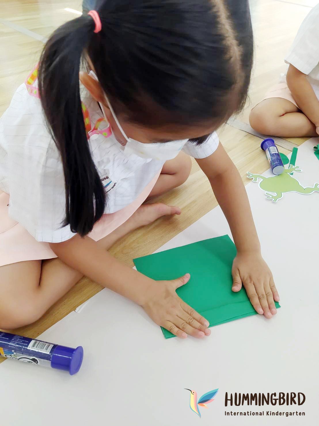 A little girl wearing a mask is sitting on the floor making a frog out of green paper.