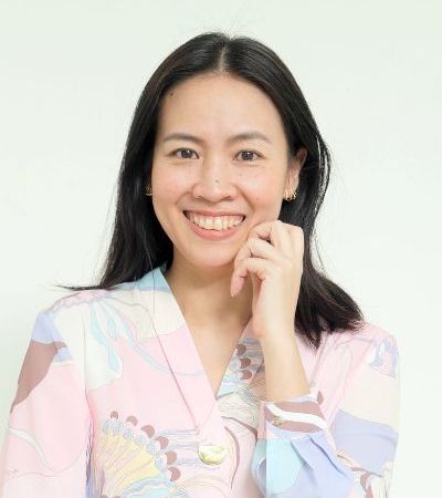 A woman in a pink jacket is smiling and touching her face.